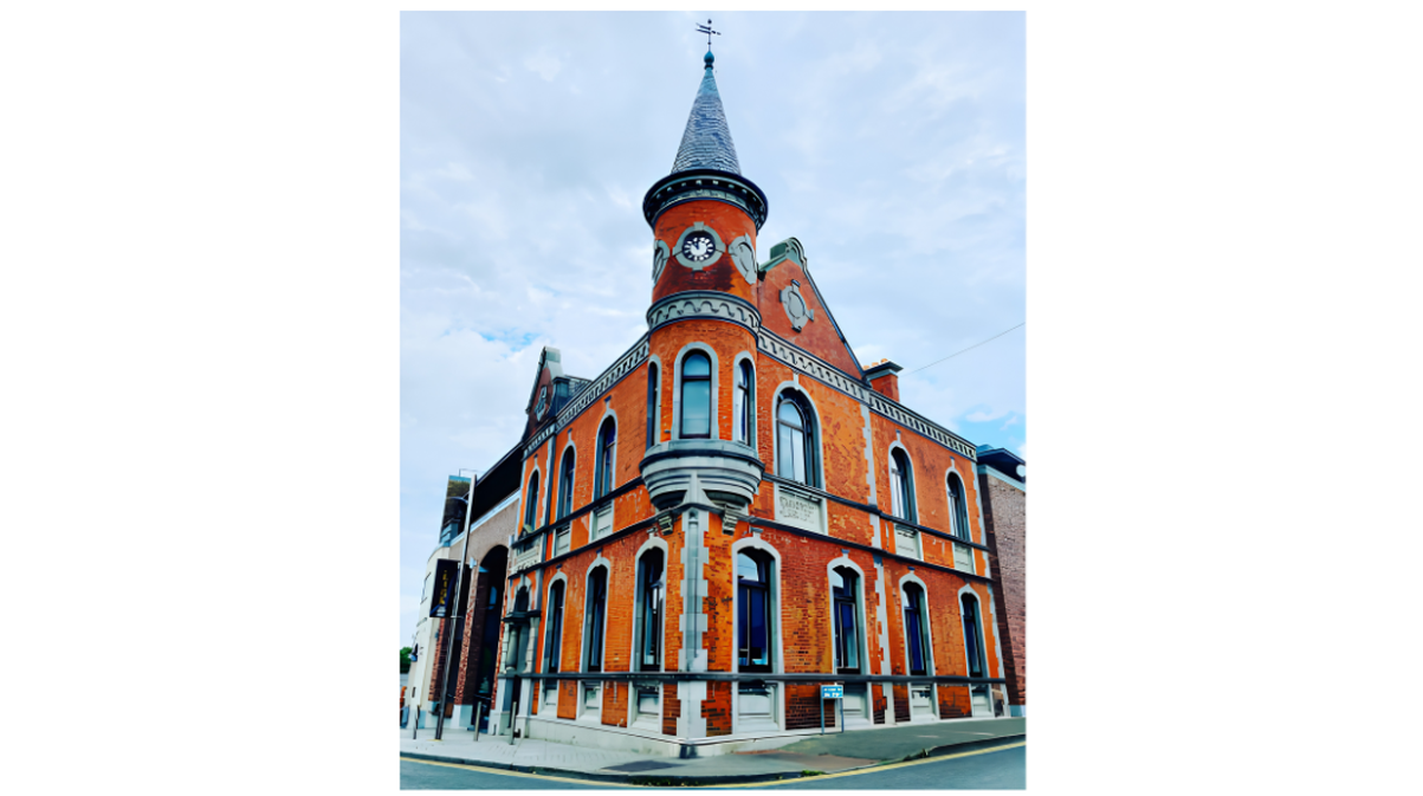 Red brick building with a tall spire and clock, featuring arched windows and decorative stonework, set on a street corner under a cloudy sky.